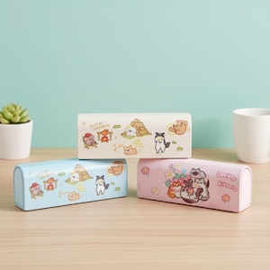 Può includere: Tre custodie rettangolari con bordi arrotondati. Una è azzurra, una bianco sporco e una rosa. Ogni custodia presenta un design a cartoni animati di animali che campeggiano o gattini con fiori. La custodia bianco sporco ha il testo "Let's go camping".