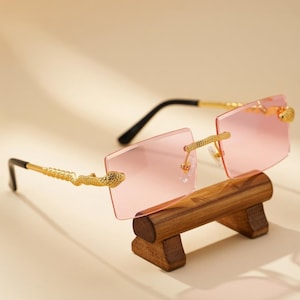 May include: Rimless sunglasses featuring rectangular light pink lenses and gold-tone metal temples. The temples are designed with a textured snake motif that connects to the lens corners. The earpieces are finished with black plastic tips.