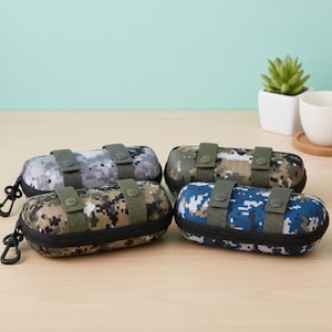 May include: Four oval-shaped eyeglass cases with camouflage patterns in grey, green, and blue. Each case has a black zip, green straps, and a black carabiner clip. They are displayed on a light brown surface.