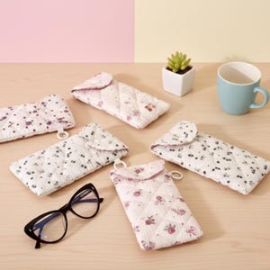 May include: Several quilted eyeglass cases in white with a floral pattern and a pink interior. A pair of black eyeglasses is also present. A blue mug and a small potted succulent are in the background.