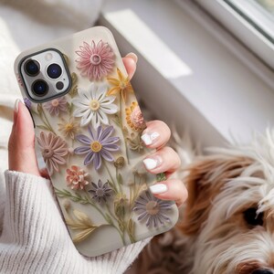 May include: A beige phone case featuring a 3D floral design with various pastel-colored flowers, including pink, white, yellow, and purple. The case has a glossy finish and a camera cutout. The phone case is a stylish accessory.