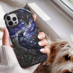 May include: A phone case featuring a stained glass style illustration of a black raven perched on an oak branch. The design includes deep blue and purple hues, a crescent moon, oak leaves, acorns, and dark roses against a gothic architectural backdrop.