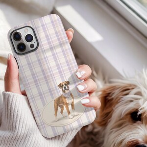 May include: A phone case with a plaid pattern in shades of lavender and cream. The case features a Boxer dog illustration wearing a bandana. The phone case is held in a person's hand.