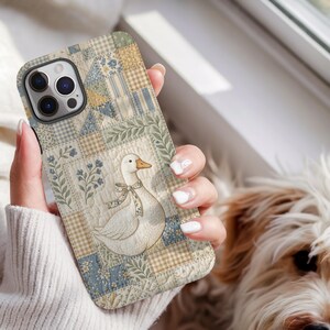 May include: A phone case featuring a quilted patchwork design in shades of blue, beige, and cream. The pattern includes squares with floral motifs, gingham checks, and a central illustration of a white goose wearing a small patterned neck ribbon.