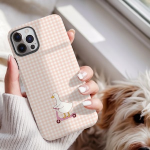 May include: A phone case with a pink and white gingham design. The case has a cartoon duck on a pink scooter. The phone case is held in a person's hand.
