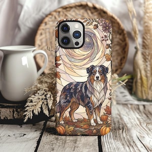 Peut inclure: Une coque de smartphone ornée d'une illustration style vitrail représentant un berger australien debout parmi des feuilles d'automne et des citrouilles. Le fond montre un ciel lunaire tourbillonnant avec des motifs floraux dans des tons marron, beige et crème.