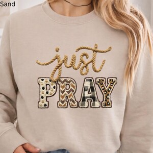 Può includere: Felpa color sabbia con le parole ricamate "just PRAY". La parola "just" è in un carattere a forma di corda e "PRAY" è in un design patchwork con pois, chevron, plaid e stampa leopardata.