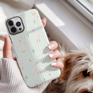 May include: A phone case with a light green and white gingham design, decorated with illustrations of white geese wearing blue bows. The case is held in a person's hand, with a dog in the background.