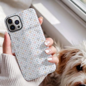 May include: A phone case featuring a light blue and white gingham pattern, adorned with tiny pink, yellow, and blue floral designs. The case is held in a hand, with a dog visible in the background.