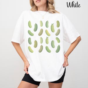 May include: White t-shirt featuring a pattern of green pickle illustrations. The pickles vary in shades of green and are scattered across the shirt. The word "White" is printed in black at the top right corner.