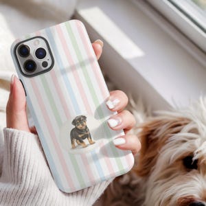 Peut inclure: Coque de téléphone à rayures pastel avec un motif de chiot. L'étui présente des rayures verticales roses, vertes et bleues. Un autocollant rond avec une illustration de chiot Rottweiler est centré sur l'étui.