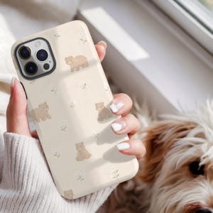 May include: A beige phone case featuring a repeating pattern of small brown bears and white floral accents. The case has a glossy finish and is designed to fit a smartphone. The phone case is held in a person's hand.