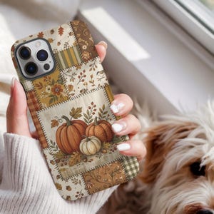 May include: A smartphone case featuring a patchwork design with autumn-themed illustrations. The pattern includes orange and white pumpkins, brown leaves, berries, acorns, and plaid fabric textures in shades of brown, tan, and muted green.