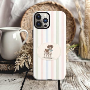 May include: A smartphone case featuring a vertical striped pattern in pastel pink, blue, and green. Centered on the case is an oval-shaped emblem containing an illustration of a brown and white spotted puppy wearing a light blue bow tie.