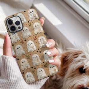 May include: A tan smartphone case featuring a repeating pattern of cream-coloured ghost figures with stitched details, small brown and green embroidered leaf accents, and tiny black stars scattered across the surface of the protective cover.