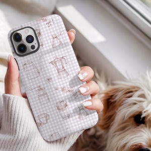 May include: A phone case with a light purple and white gingham pattern, featuring brown line drawings of coffee-making equipment. The case includes images of a coffee maker, grinder, and coffee beans. The phone case is held by a person.