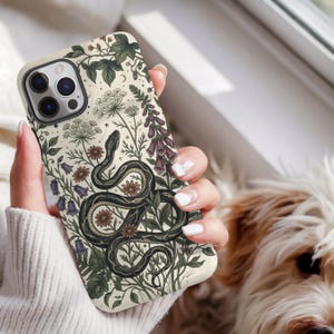 May include: A phone case with a cream background featuring a detailed illustration of a black snake coiled among various botanical elements including green leaves, purple bell-shaped flowers, tall pinkish-purple stalks, and small brown flowers.