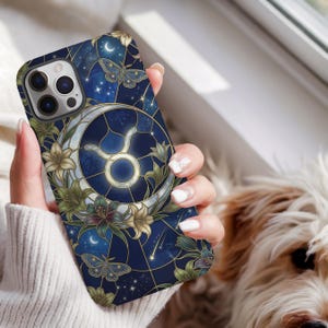 May include: A dark blue smartphone case featuring a stained-glass style design with a central Taurus zodiac symbol, a crescent moon, blooming lilies, and moths. The artwork includes thin gold lines, scattered stars, and a deep navy background.