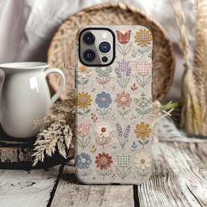 May include: A smartphone case with a light grey background featuring a repeating pattern of embroidered-style flowers and tulips in various colours including blue, yellow, red, and purple, with checkered and plaid fabric textures on the petals.