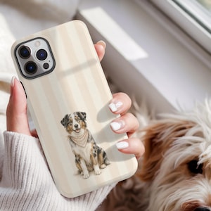Könnte beinhalten: Eine Smartphone-Hülle mit vertikalem Streifenmuster in Creme und Beige sowie einer zentralen Illustration eines sitzenden Australian Shepherd Hundes mit einem hellen Halstuch. Die Hülle hat einen schwarzen Kameraausschnitt oben links.