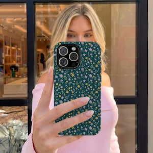 May include: A teal phone case with a floral pattern of small yellow and pink flowers. The case has a camera cutout with three lenses and a flash. The phone case is being held up in front of a person.