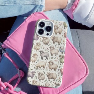 May include: A phone case with a repeating pattern of sheep in various colors and poses. The case is resting on a pink purse. The sheep are depicted in a watercolor style, with details like wool texture and floral accents.