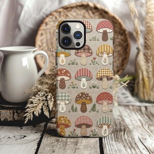 May include: A smartphone case featuring a repeating pattern of various mushrooms on a light beige background. The mushrooms display diverse textures, including polka dots, plaid, and floral prints in shades of red, yellow, brown, and green.