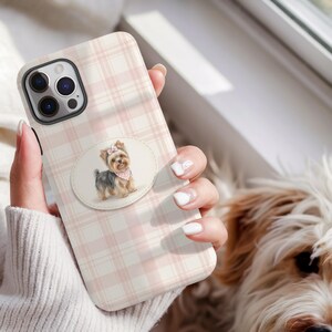 Peut inclure: Coque de téléphone à motif à carreaux roses et blancs. La coque présente un motif circulaire avec une illustration à l'aquarelle d'un Yorkshire Terrier portant un nœud et un bandana roses. La coque est tenue par une personne.