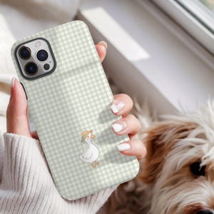 May include: A phone case with a light green and white gingham pattern. The case features a cartoon goose wearing a straw hat. The phone case is held in a person's hand.