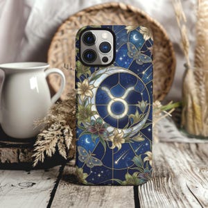 May include: A smartphone case featuring a dark blue stained glass design with a central gold Taurus zodiac symbol, a crescent moon, lilies, moths, and stars. The pattern includes geometric lines and celestial elements in shades of blue and gold.