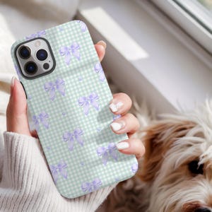May include: A phone case with a light green and white gingham design, adorned with lavender bow illustrations. The case, held in a hand, wraps around the phone's back and sides, offering protection. The design features repeating bow patterns.