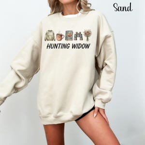 May include: A sand-colored crewneck sweatshirt featuring a graphic print of a sweater, a coffee mug, a book with a leaf, binoculars, and a bouquet of flowers. Below the icons, the text HUNTING WIDOW is printed in a bold, black, sans-serif font.