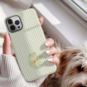 May include: A phone case with a light green and white gingham pattern. A cartoon frog playing a ukulele is featured on the case. The phone case is held by a person.