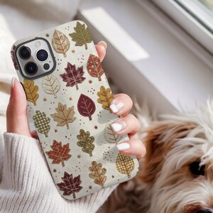 May include: A smartphone case featuring a cream-coloured background with a repeating pattern of various autumn leaves in shades of burgundy, olive green, mustard yellow, and brown. The leaves display stitched details and plaid or solid textures.