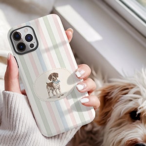 May include: A smartphone case with a pastel striped design in pink, green, and blue. The case has an oval illustration of a brown and white dog wearing a blue bow tie. The case is held by a person.