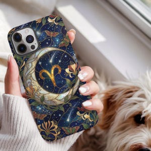 Puede incluir: Una funda para smartphone con un fondo de cielo nocturno estrellado en azul oscuro, una gran luna creciente, el símbolo dorado del zodiaco Aries e ilustraciones detalladas de flores y polillas en tonos amarillo, verde y rojo.