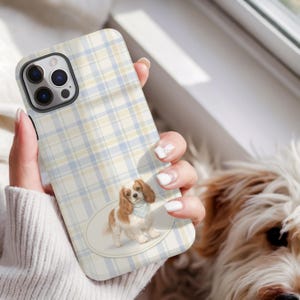 May include: A phone case with a light blue and yellow plaid pattern. The case features an illustration of a Cavalier King Charles Spaniel dog wearing a blue bandana. The phone case is held by a person.