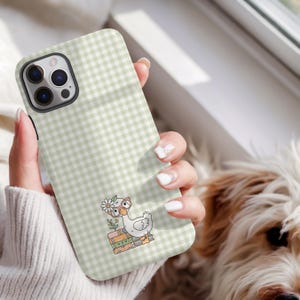 May include: A phone case with a pale green and white gingham design. The case displays a cartoon goose wearing spectacles and a flower, perched on books. The phone case is held in a person's hand.