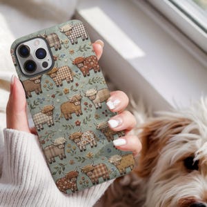 May include: A phone case featuring a repeating pattern of Highland cattle on a sage green background. The cattle are depicted in various brown and tan plaid, floral, and solid textures, interspersed with small colourful flowers and green sprigs.