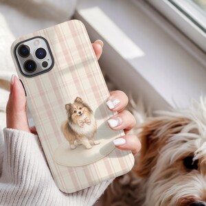 May include: A phone case with a pink and cream plaid pattern. The case features an illustration of a fluffy, brown and white dog wearing a pink bow tie. The phone case is held by a person.