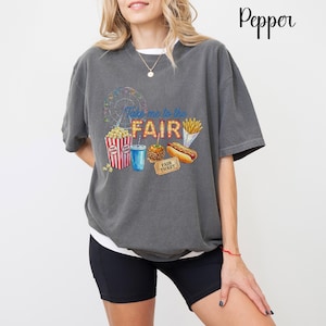 Pode incluir: T-shirt cinzento escuro com um gráfico colorido de feira. O gráfico inclui uma roda gigante, pipocas, uma bebida, uma maçã caramelizada, um cachorro-quente, batatas fritas e um bilhete. O texto "Take me to the FAIR" também está presente.