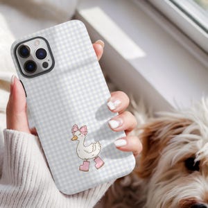 May include: A phone case with a light blue and white gingham design. The case displays a cartoon goose wearing a pink bow and pink boots. The phone case is held in a person's hand.