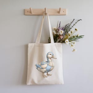 May include: A cream-colored tote bag with a detailed illustration of a duck. The duck features a patchwork design in shades of blue, tan, and white. The bag hangs from a wooden rack on a light gray wall, with a bouquet of dried flowers inside.