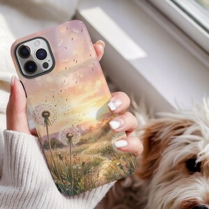 May include: A smartphone case featuring a printed illustration of a dandelion field at sunset. The design displays white dandelion seed heads blowing in the wind against a soft gradient background of pastel pink, orange, and yellow hues.