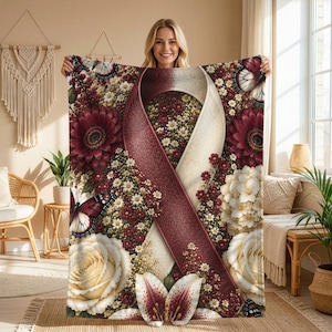 May include: A throw blanket featuring a large awareness ribbon in burgundy and cream, surrounded by a dense floral pattern of burgundy gerbera daisies, cream roses, white hydrangeas, small white flowers, and several butterflies with dark wings.