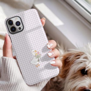 May include: A phone case with a light purple and white gingham pattern. The case features a cartoon duck holding a bouquet of flowers. The phone case is held in a person's hand.