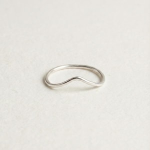 Può includere: Un anello sottile in argento lucido caratterizzato da una fascia liscia e continua con una singola punta a forma di V al centro. Il metallo ha una finitura metallica riflettente e un profilo arrotondato, creando una silhouette curva e semplice.