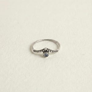 Könnte beinhalten: Ein silberner Ring mit einem dünnen, chevronförmigen Band, das am oberen Rand eine perlenartige Textur aufweist. Ein kleiner, runder, durchscheinender blaugrauer Stein ist am Mittelpunkt des Chevrons gefasst und bildet ein filigranes Schmuckstück.