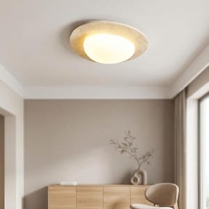 May include: A modern ceiling light fixture with a circular, textured, beige frame and a bright, white, oval-shaped light source. The light is mounted on a white ceiling. The room has neutral-toned walls and wooden furniture.