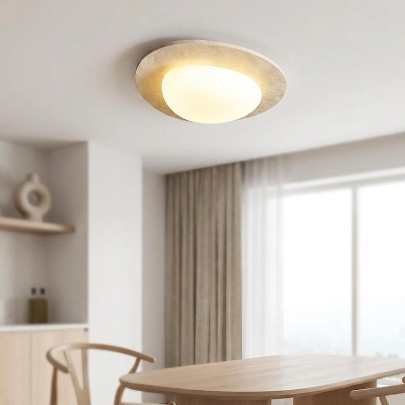 May include: A modern ceiling light fixture with a circular, textured, beige outer ring and a bright, oval-shaped, white light source. The light is mounted on a white ceiling, with a dining table and chairs in the foreground.